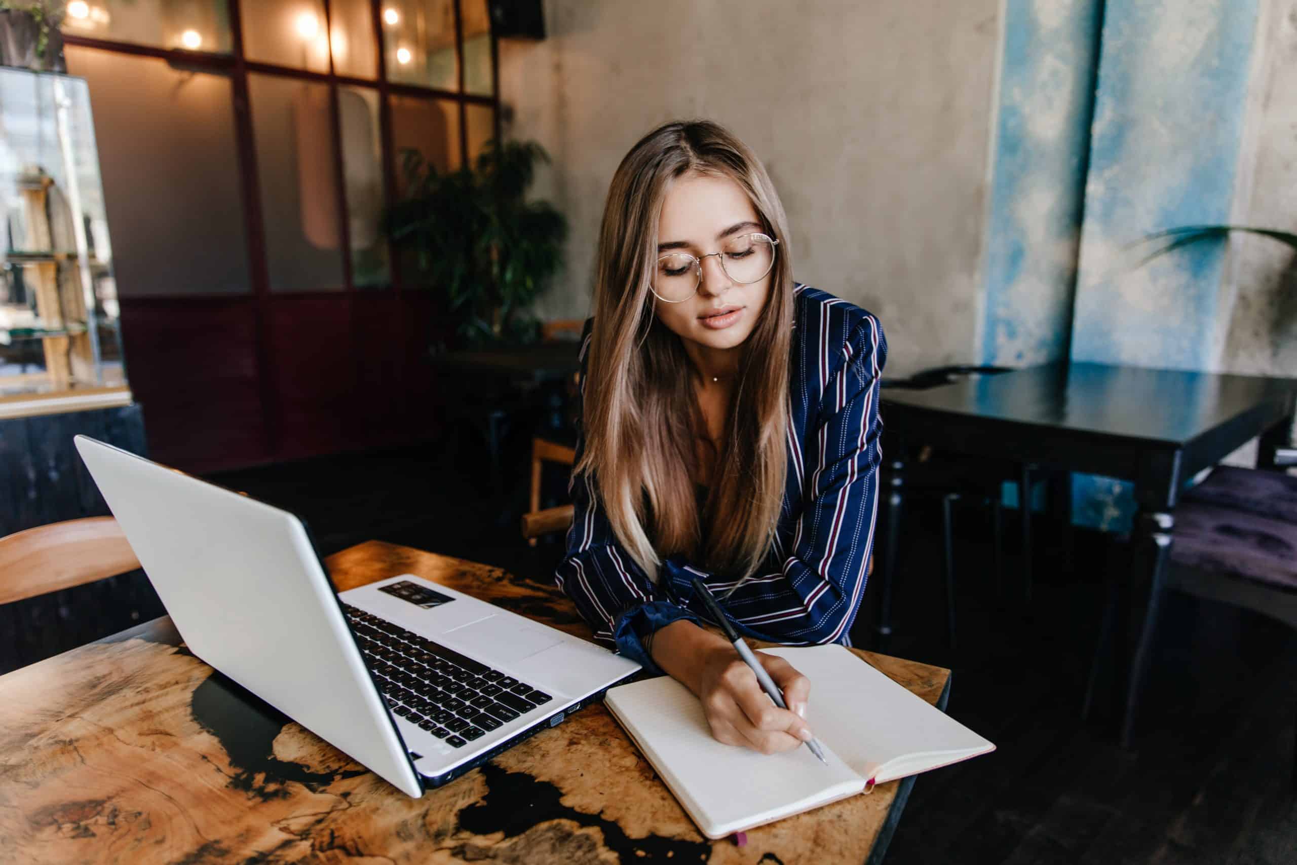 lovable girl writing something her notebook indoor shot adorable long haired woman using white laptop cafe scaled